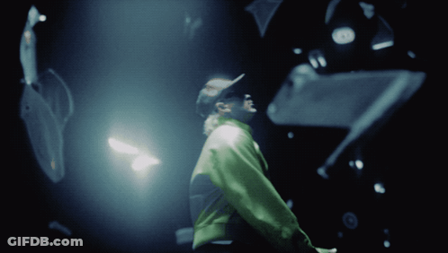 A Man In A Green Jacket Stands In A Dark Room Looking Up GIF