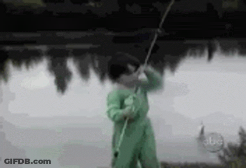 A Man In A Green Jumpsuit Is Holding A Fishing Rod In Front Of A Lake . GIF