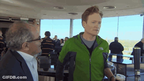A Man In A Green North Face Jacket Talks To Another Man In A Suit GIF
