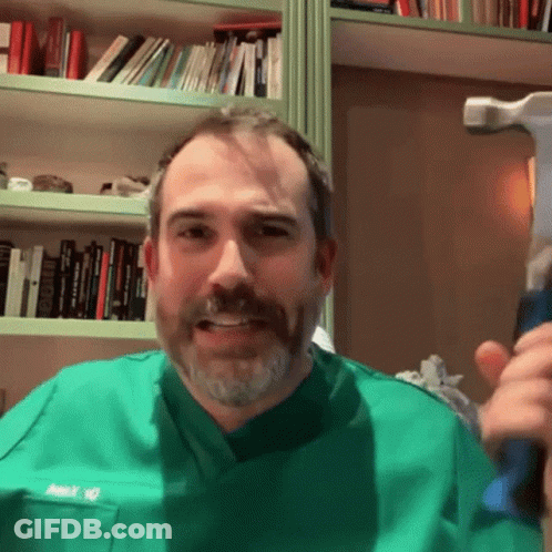 A Man In A Green Scrub Top Holds A Hammer In Front Of A Bookshelf GIF