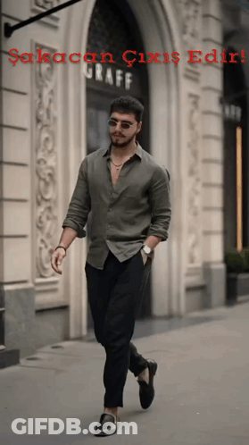 A Man In A Green Shirt And Black Pants Walks Down A Sidewalk In Front Of A Building That Says Graff GIF