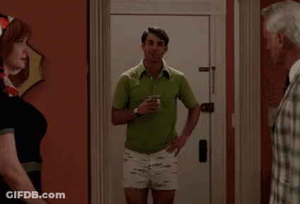 A Man In A Green Shirt And White Shorts Is Standing In Front Of A Door Holding A Cup Of Coffee . GIF