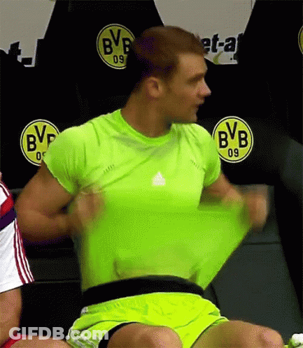 A Man In A Green Shirt And Yellow Shorts Is Taking Off His Shirt In Front Of A Bvb Logo . GIF