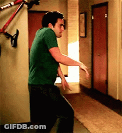 A Man In A Green Shirt Is Dancing In A Hallway With A Bike Hanging On The Wall Behind Him . GIF