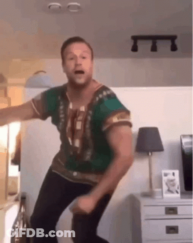 A Man In A Green Shirt Is Dancing In A Living Room With A Lamp In The Background . GIF