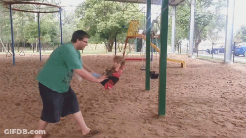 A Man In A Green Shirt Is Pushing A Child On A Swing At A Playground GIF