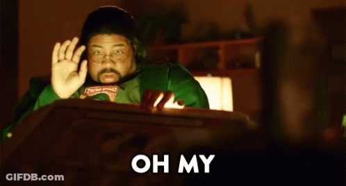 A Man In A Green Shirt Is Sitting At A Table In Front Of A Computer Screen And Waving . GIF