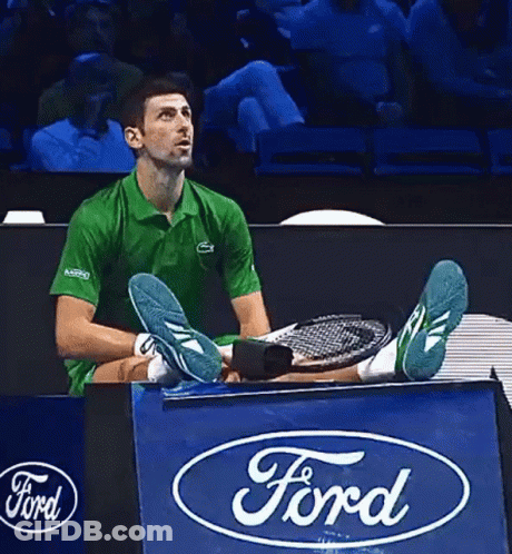 A Man In A Green Shirt Is Sitting On A Ford Sign Holding A Tennis Racquet GIF