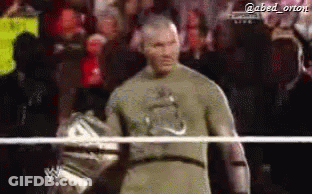 A Man In A Green Shirt Is Standing In A Wrestling Ring With A Crowd Behind Him . GIF