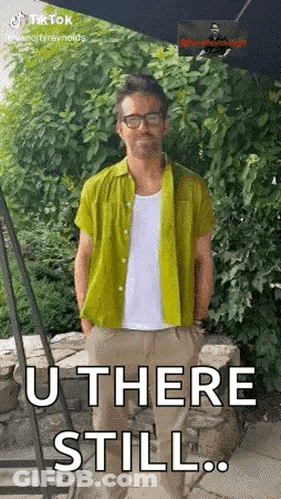 A Man In A Green Shirt Is Standing In Front Of A Stone Wall With His Hands In His Pockets . GIF