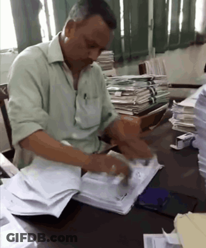 A Man In A Green Shirt Sits At A Desk With A Stack Of Papers GIF