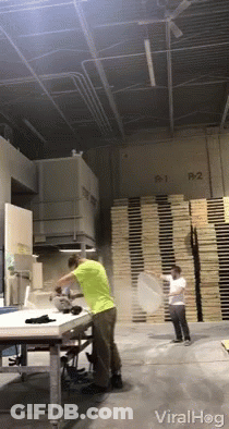 A Man In A Green Shirt Throws A Pizza In A Warehouse With A Stack Of Wooden Pallets In The Background GIF