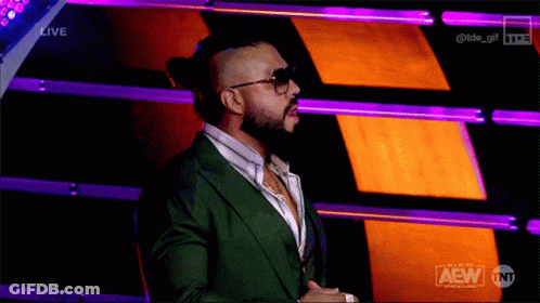 A Man In A Green Suit And Sunglasses Stands In Front Of A Purple And Orange Background That Says Aew On It GIF