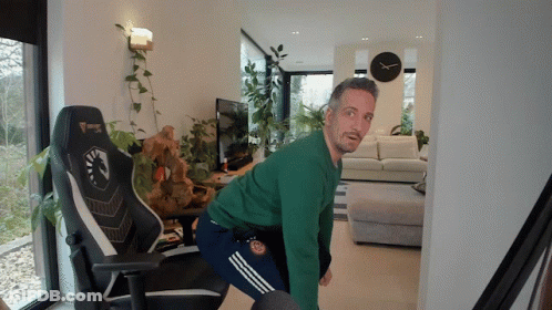 A Man In A Green Sweater Is Squatting In Front Of A Gaming Chair GIF