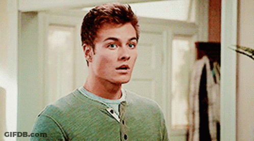 A Man In A Green Sweater Is Standing In Front Of A Door Looking Surprised . GIF
