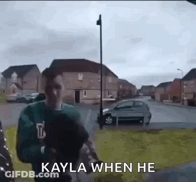 A Man In A Green Sweater Is Walking Down A Street With The Words `` Kayla When He '' Written On It . GIF