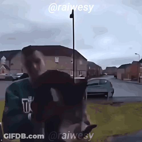 A Man In A Green Sweatshirt Is Standing In Front Of A Street With The Name @Raiwesy On The Bottom GIF