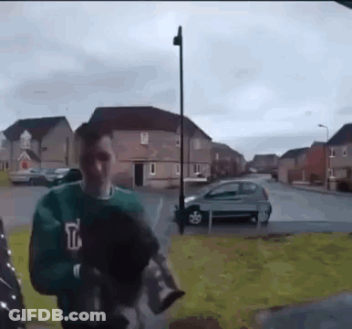 A Man In A Green Sweatshirt With The Letter T On It Is Walking Down A Street . GIF