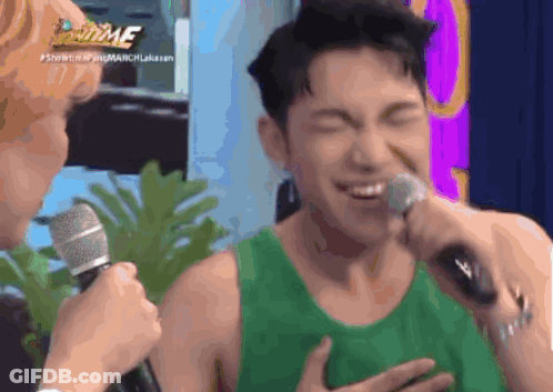 A Man In A Green Tank Top Is Singing Into A Microphone While Another Man Looks On .. GIF