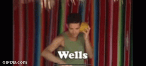 A Man In A Green Tank Top Is Standing In Front Of A Colorful Curtain Holding A Yellow Object In His Hand . GIF