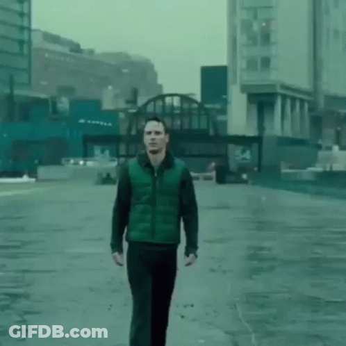 A Man In A Green Vest And Black Pants Is Walking Down A Wet Street In A City . GIF