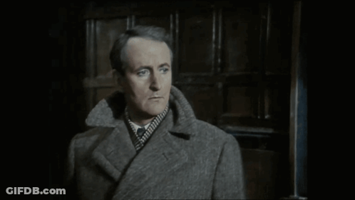 A Man In A Grey Coat Is Standing In Front Of A Door GIF