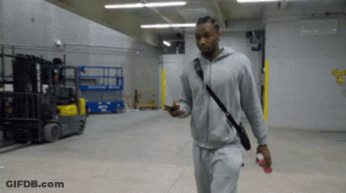 A Man In A Grey Hoodie Is Walking In A Warehouse GIF