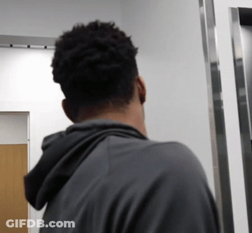 A Man In A Grey Hoodie Stands In A Hallway GIF