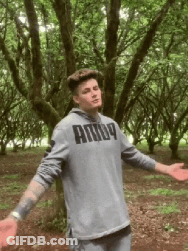 A Man In A Grey Puma Hoodie Stands In A Forest With His Arms Outstretched GIF