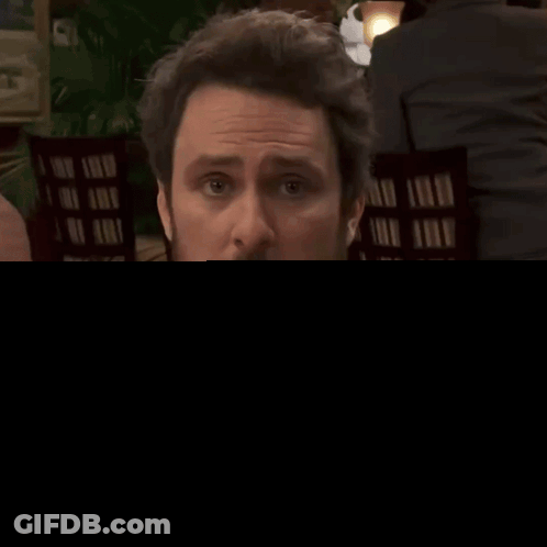 A Man In A Grey Shirt And Tie Is Sitting At A Table With A Woman Behind Him GIF
