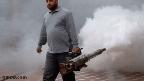 A Man In A Grey Shirt Is Holding A Machine That Is Shooting Smoke GIF