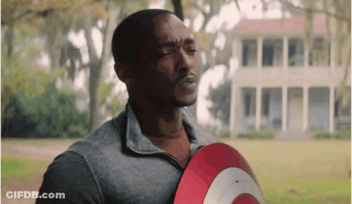 A Man In A Grey Shirt Is Holding A Red And White Shield In Front Of A House . GIF