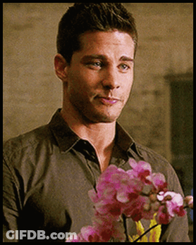 A Man In A Grey Shirt Is Holding Pink Flowers In His Hand GIF