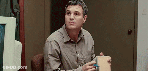 A Man In A Grey Shirt Is Sitting In A Chair Holding A Box And A Pen . GIF