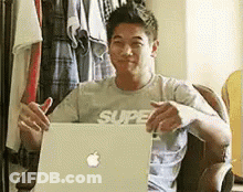 A Man In A Grey Shirt Is Sitting In Front Of An Apple Laptop . GIF