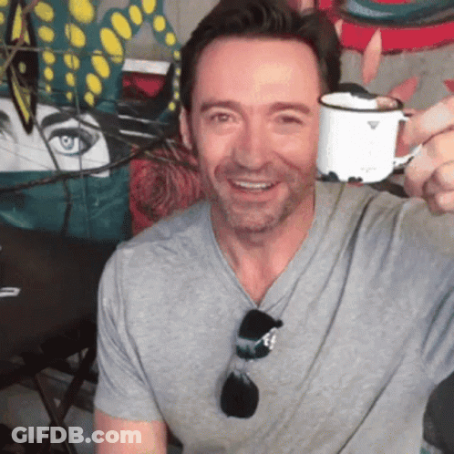 A Man In A Grey Shirt Is Smiling And Holding A White Cup GIF
