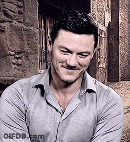 A Man In A Grey Shirt Is Smiling With His Arms Crossed In Front Of A Stone Wall GIF