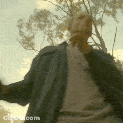 A Man In A Grey Shirt Is Standing In Front Of A Tree GIF