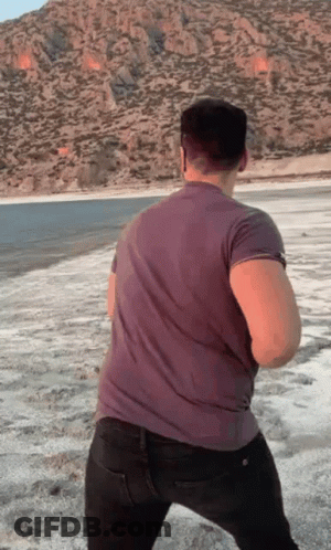 A Man In A Grey Shirt Is Standing On A Beach With Mountains In The Background GIF