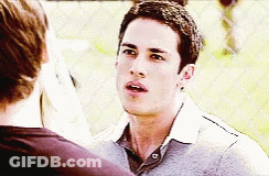A Man In A Grey Shirt Is Talking To Another Man Behind A Chain Link Fence GIF