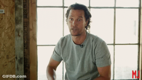 A Man In A Grey Shirt Stands In Front Of A Window With The Letter M On The Bottom GIF