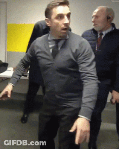 A Man In A Grey Sweater And Tie Stands In A Room With Other Men GIF