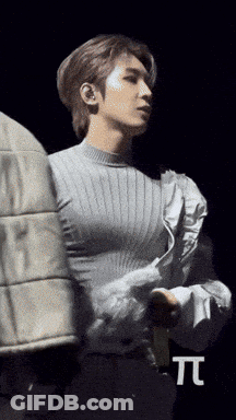 A Man In A Grey Sweater Is Standing In Front Of A Black Background With The Letter T. GIF