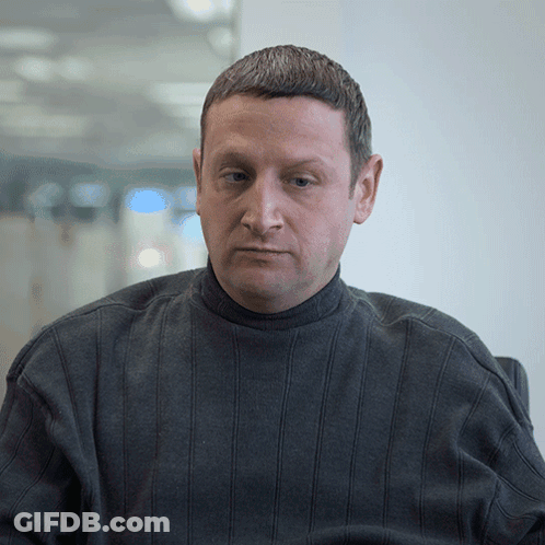 A Man In A Grey Sweater Looks At The Camera With A Serious Look On His Face GIF
