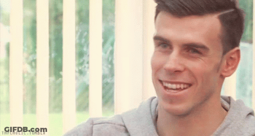 A Man In A Grey Sweatshirt Is Smiling In Front Of A Window With A Striped Blind Behind Him GIF