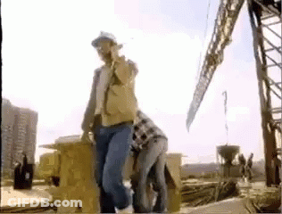 A Man In A Hard Hat Is Standing Next To A Woman In A Plaid Shirt On A Construction Site . GIF