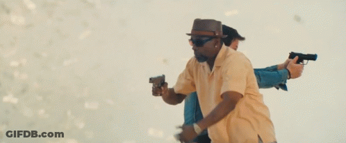 A Man In A Hat And Sunglasses Is Holding Two Guns In Front Of A Woman . GIF