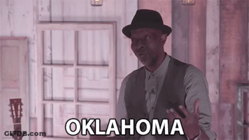 A Man In A Hat And Vest Is Standing In Front Of A Window And Holding A Guitar . GIF