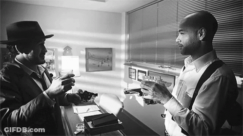 A Man In A Hat Holds A Gun Next To Another Man Who Holds A Gun GIF