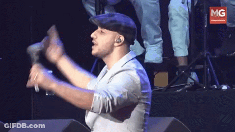 A Man In A Hat Holds A Microphone In Front Of A Banner That Says Mg GIF